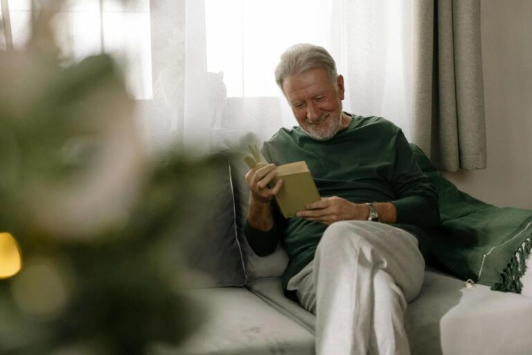 An elderly person reading a book at home, focusing on the text with glasses.