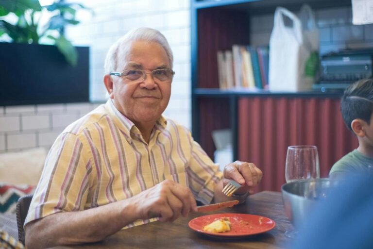 A retired man enjoying a meal at home, smiling at the camera.