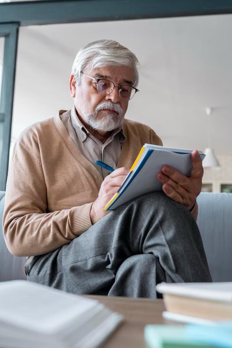 An elderly man sitting on the couch reviewing a Medicare enrollment checklist with a notebook.