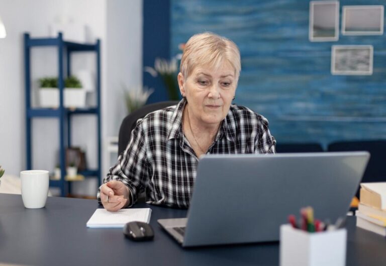 An elderly woman thoughtfully reviewing documents on her laptop.