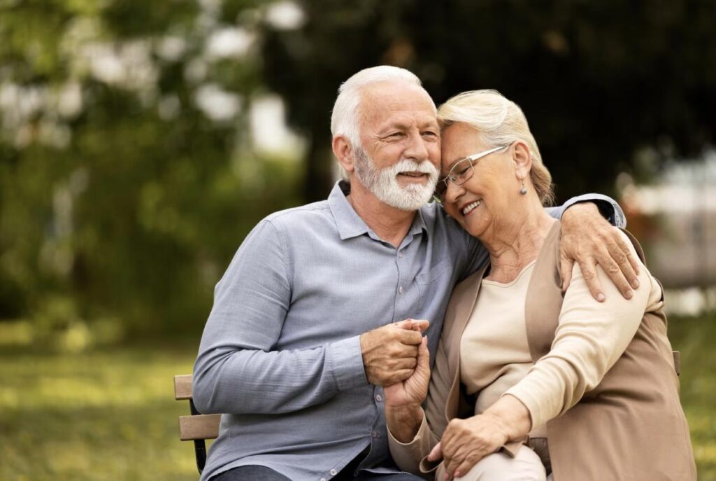 An elderly couple sitting on a park bench, smiling and holding hands.