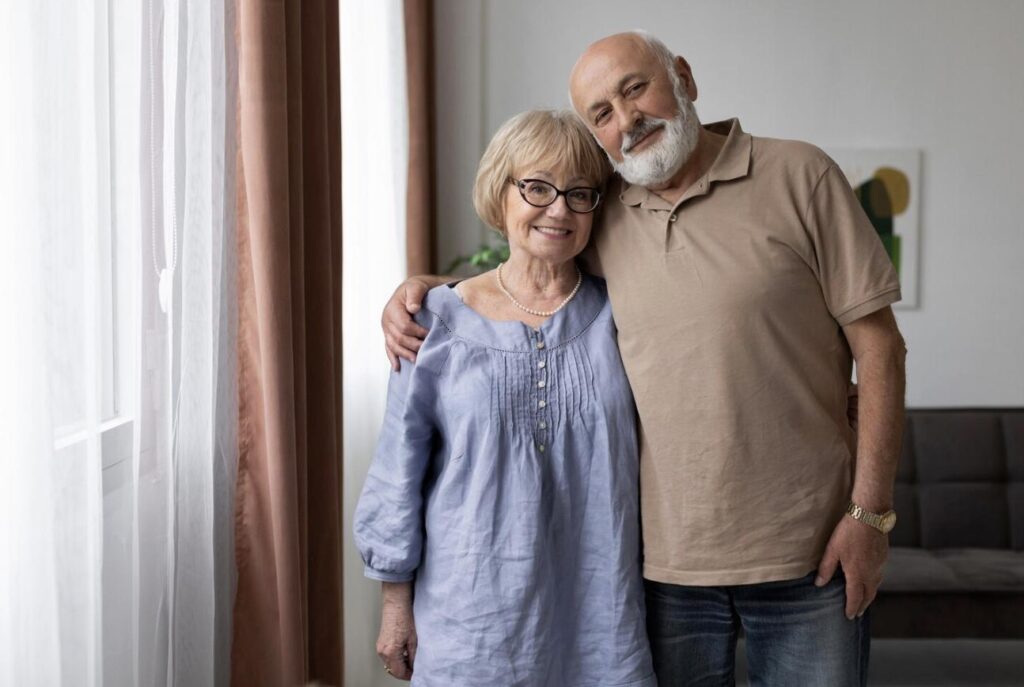 An elderly couple smiling and posing together in their home.