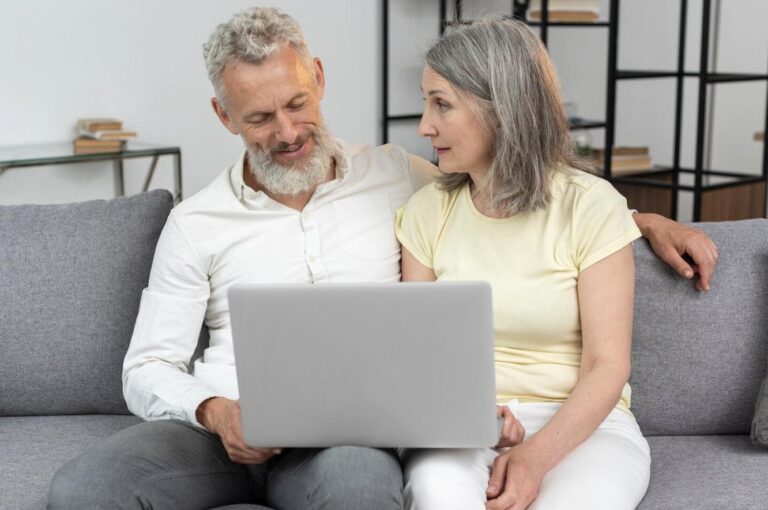 An elderly couple at home on a couch, looking at updates on a laptop.