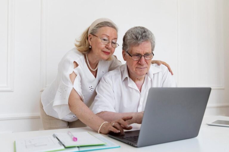 An elderly couple sitting at a wooden table, looking at a laptop screen together.