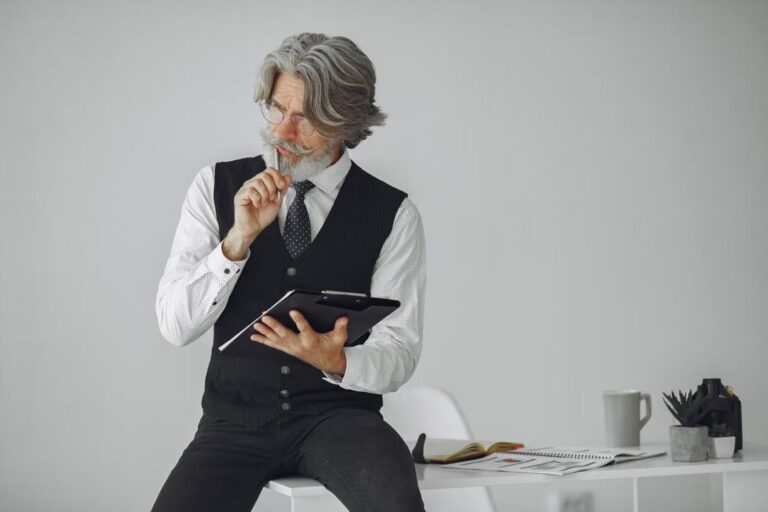 A thoughtful senior man with gray hair sits at a desk holding a tablet in a modern office.