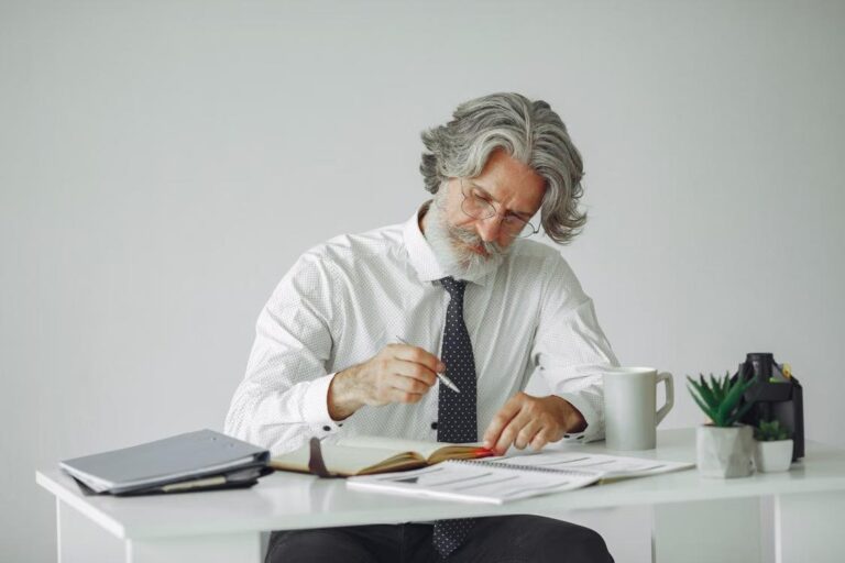 A thoughtful senior man with gray hair sits at a desk going through documents in a modern office.