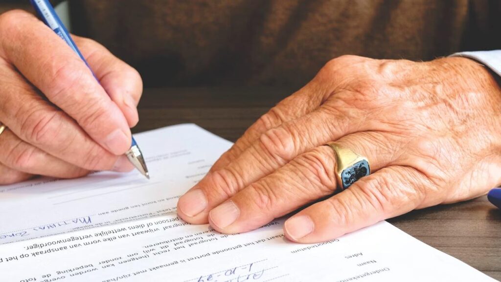 An elderly person's hand signs a document with a pen at a desk.
