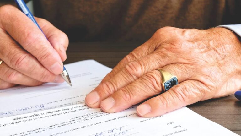 An elderly person's hand signs a document with a pen at a desk.