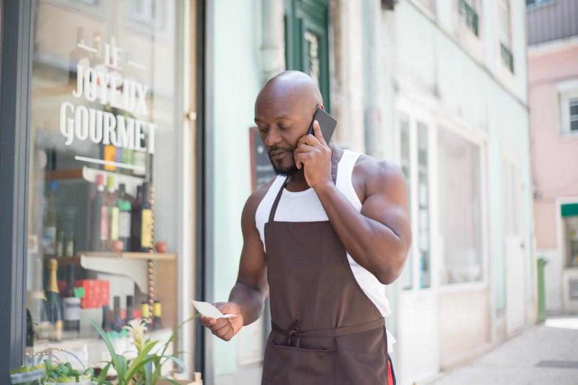 A man reads a note while talking on the phone outdoors.