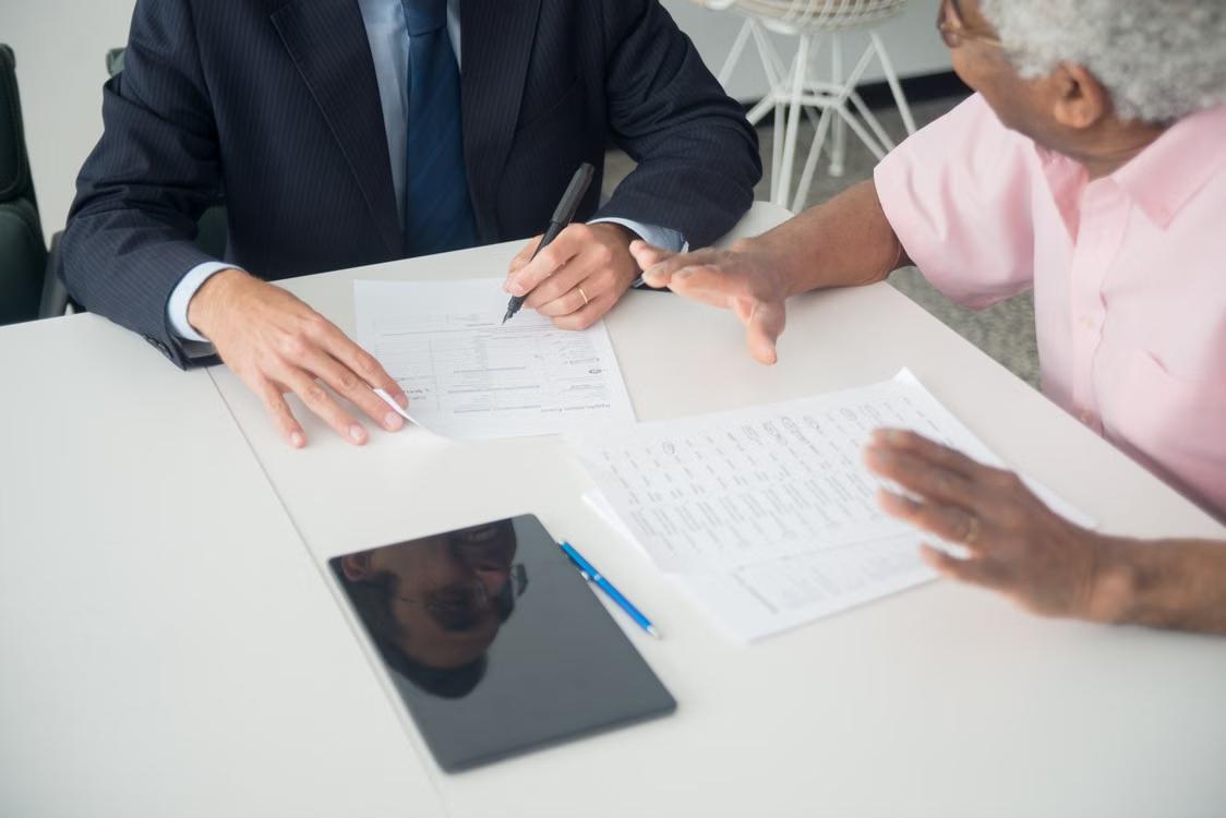 An elderly man consults with an agent while reviewing documents at a table.
