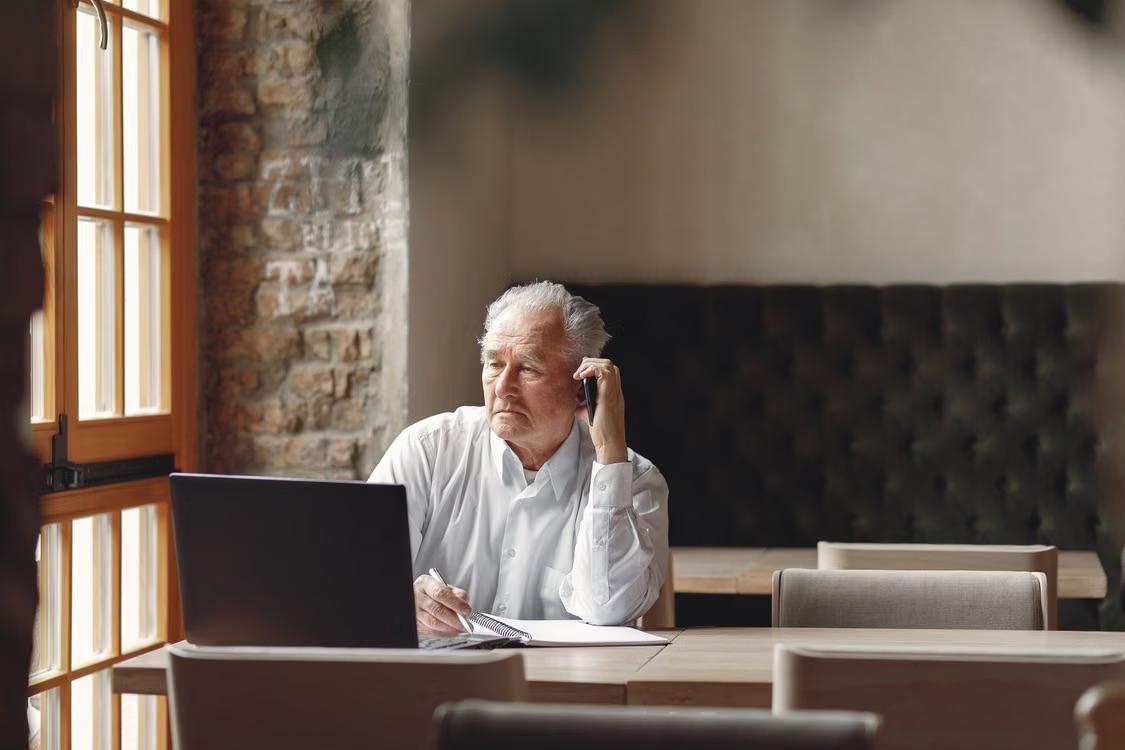 A senior man speaks on his smartphone while seated at a desk.